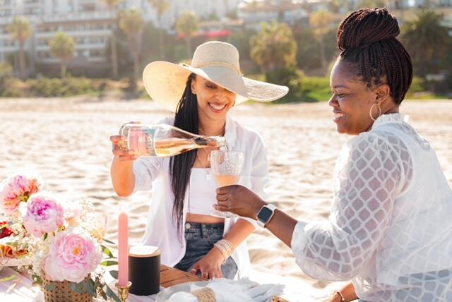 Two women sitting on a blanket on a bach. The woman on the left is pouring wine into the glass of the woman on the right 