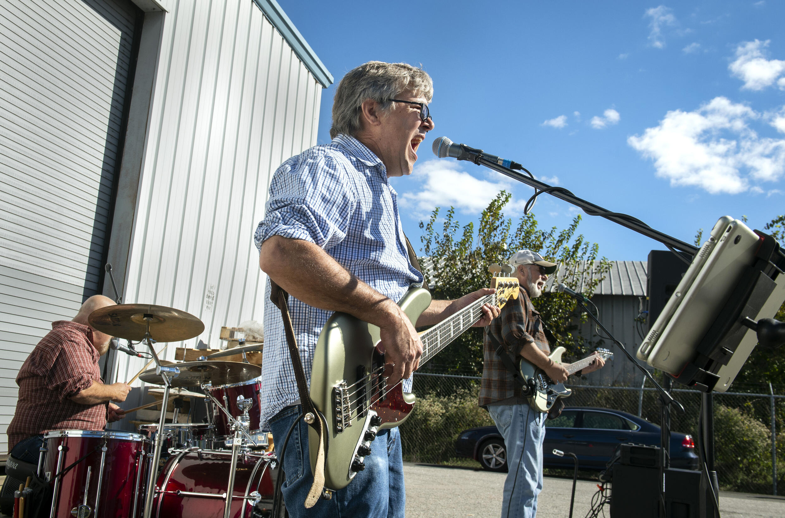 A man playing a voice guitar and singing into a microphone. Behind him is a man playing on drums and a man playing the guitar. 