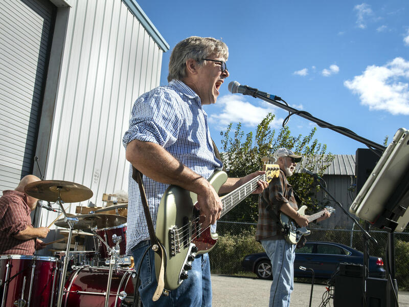 A man playing a voice guitar and singing into a microphone. Behind him is a man playing on drums and a man playing the guitar. 