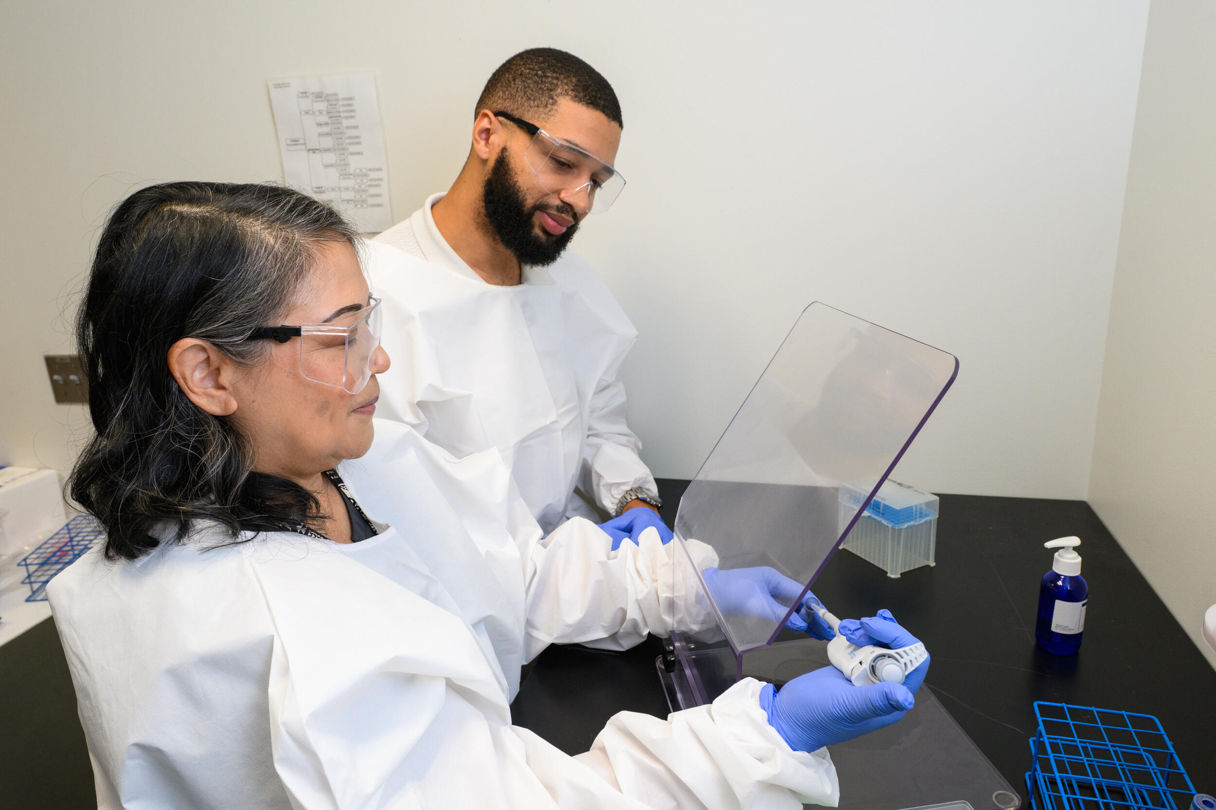 A photo of a man and woman wearing safety goggles and blue latex gloves sitting at a desk. 
