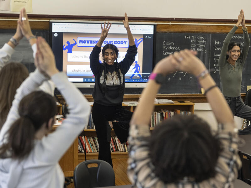 Two students at the front of a classroom balance on one leg and hold their hands above their head. Students with their backs to the camera do the same in the foreground.