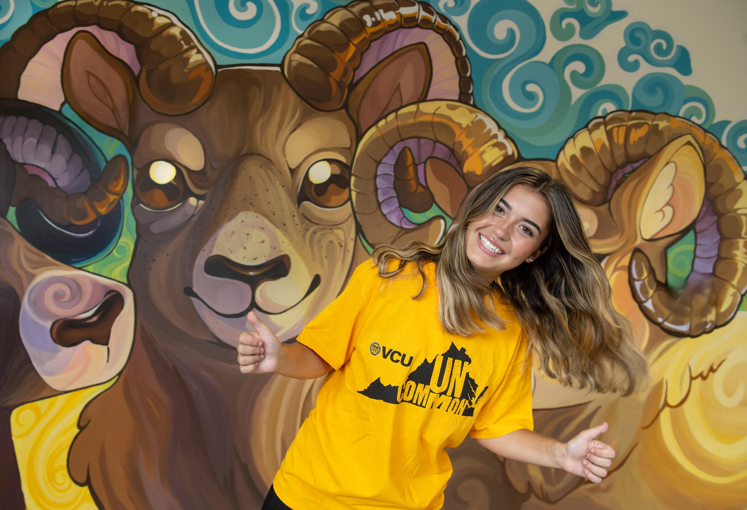 A photo of a woman from the waist up. She is leaning to the right with both of her hands making thumbs up gestures. She is wearing a yellow t-shirt with the shape of Virginia printed on it. Inside the state's silhouette is yellow text that reads \"UNCOMMON.\" Behind the woman is a mural of three rams. 