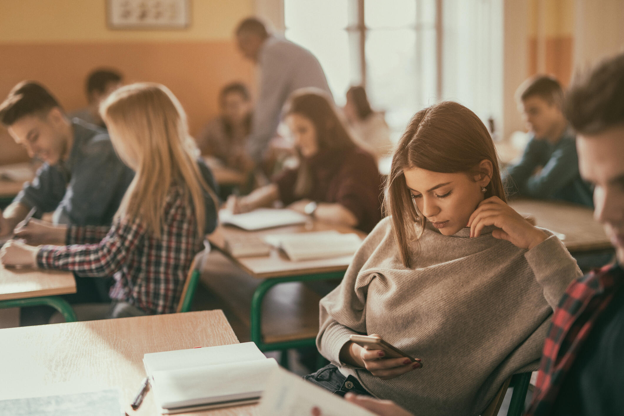 A photo of a classroom full of student sitting at desks. While most of the students are looking at books or writing, one girl is looking at her cell phone. 