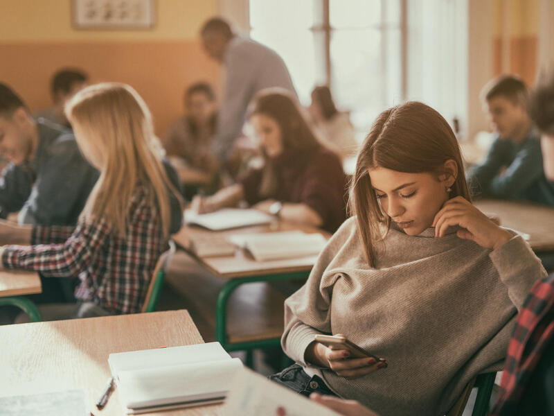 A photo of a classroom full of student sitting at desks. While most of the students are looking at books or writing, one girl is looking at her cell phone. 