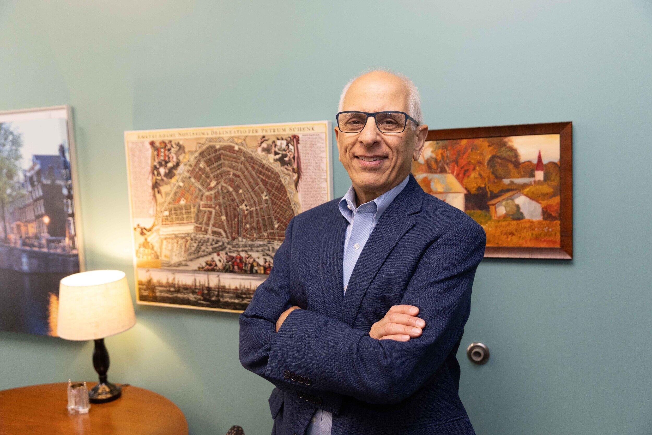 A photo of a man fromt he waist up. He is standing against a wall with his arms crossed against his chest