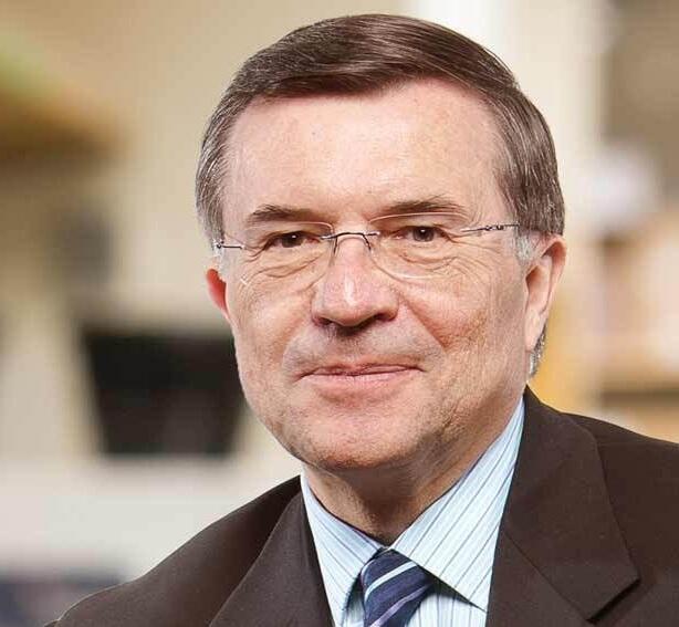 A photo portrait of a man wearing a dark blue and white stripped tie, a dark gray suit jacket, a white shirt with thin blue stripes, and glasses. 