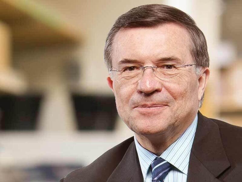 A photo portrait of a man wearing a dark blue and white stripped tie, a dark gray suit jacket, a white shirt with thin blue stripes, and glasses. 