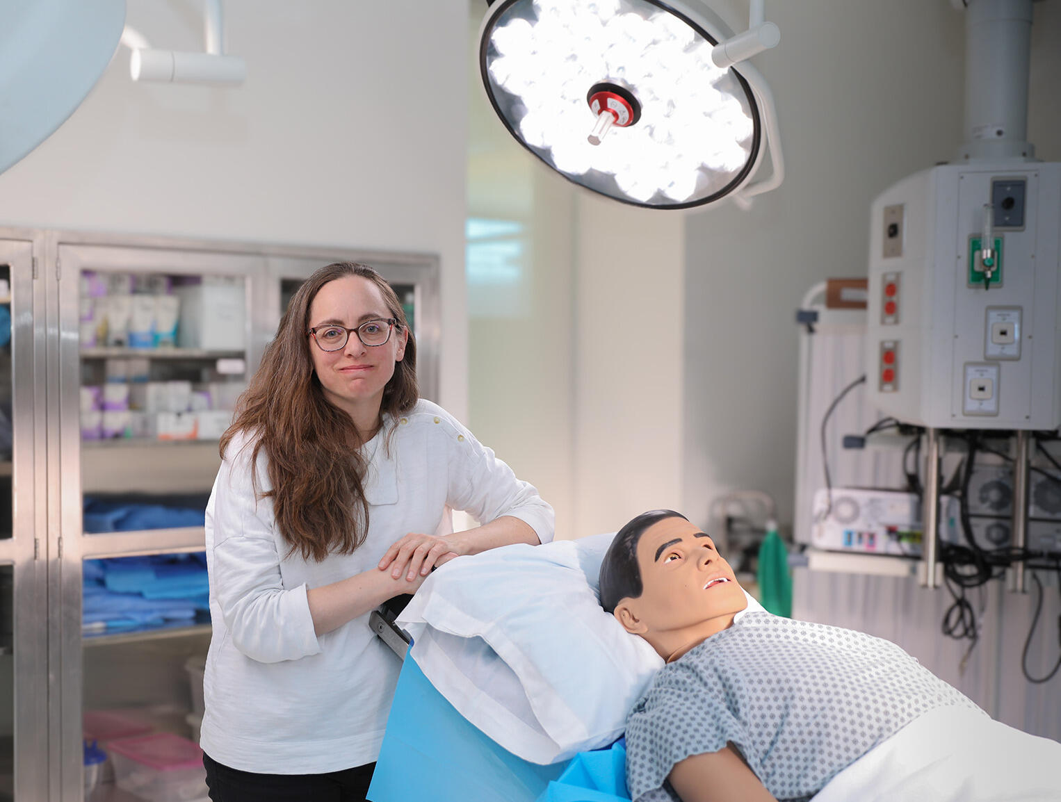 A photo of a woman lenaing on the back of a surgical table in a medical room. On the table is a dummy dressed in a hospital gown. 