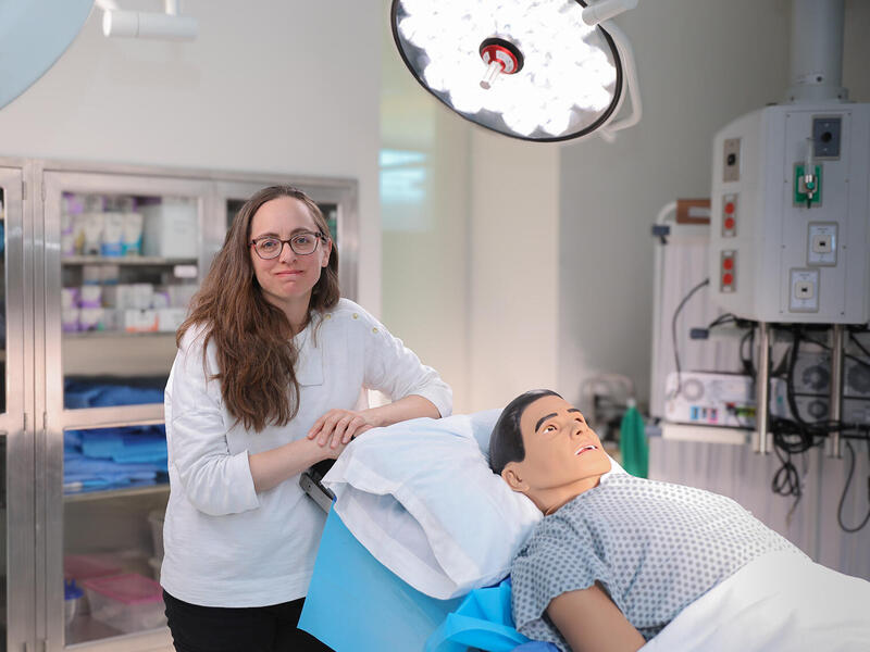 A photo of a woman lenaing on the back of a surgical table in a medical room. On the table is a dummy dressed in a hospital gown. 