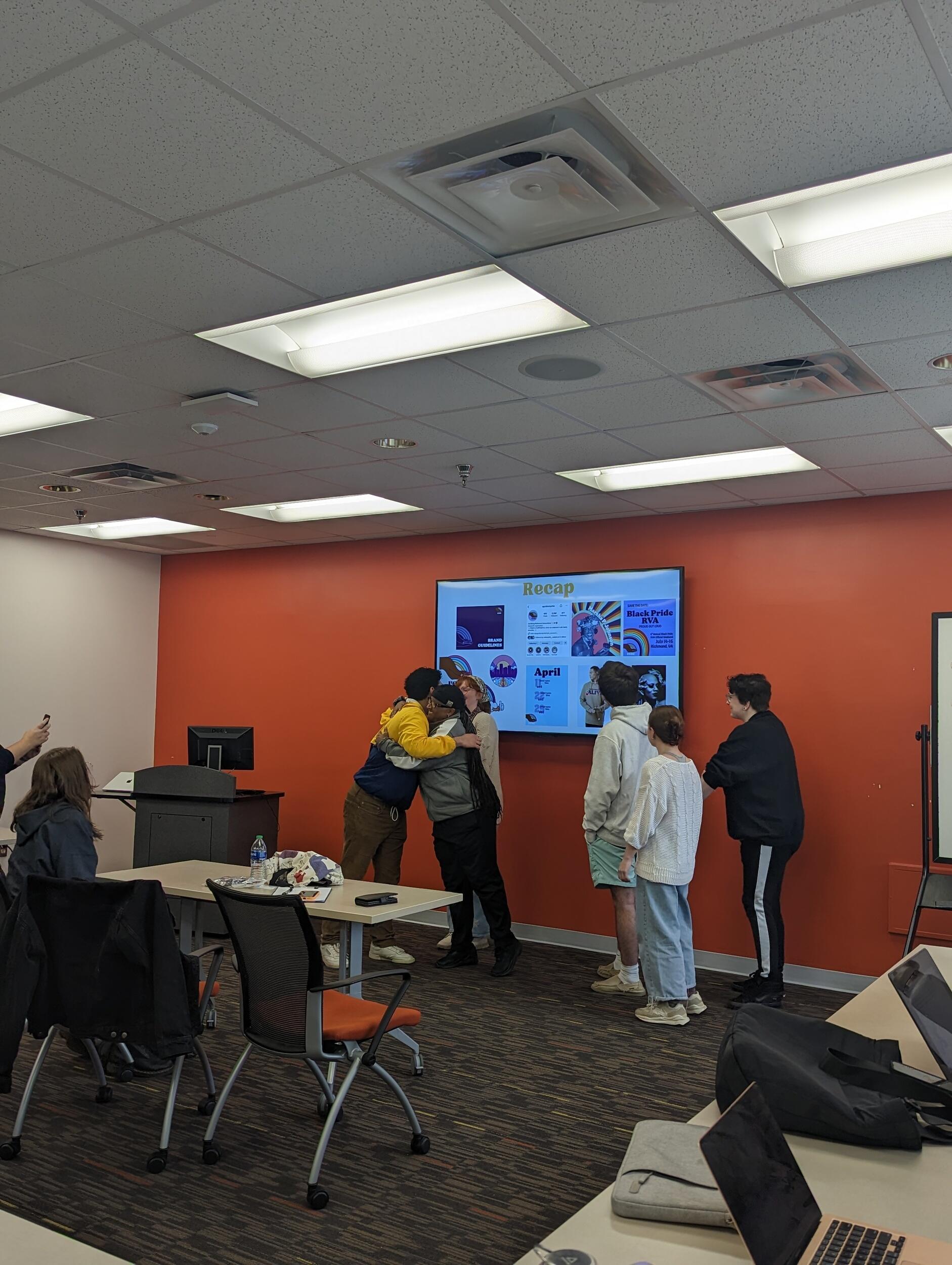 Three people are hugging in the front of a room and three other people are standing to their left. 