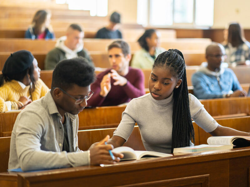 A photo of a row of lecture seats in a college classroom. There are students scattered throughout the rows. In the front is a Black man and woman who are reading off of the same book. The woman is speaking and the man is holding a pencil and one page of the book. 