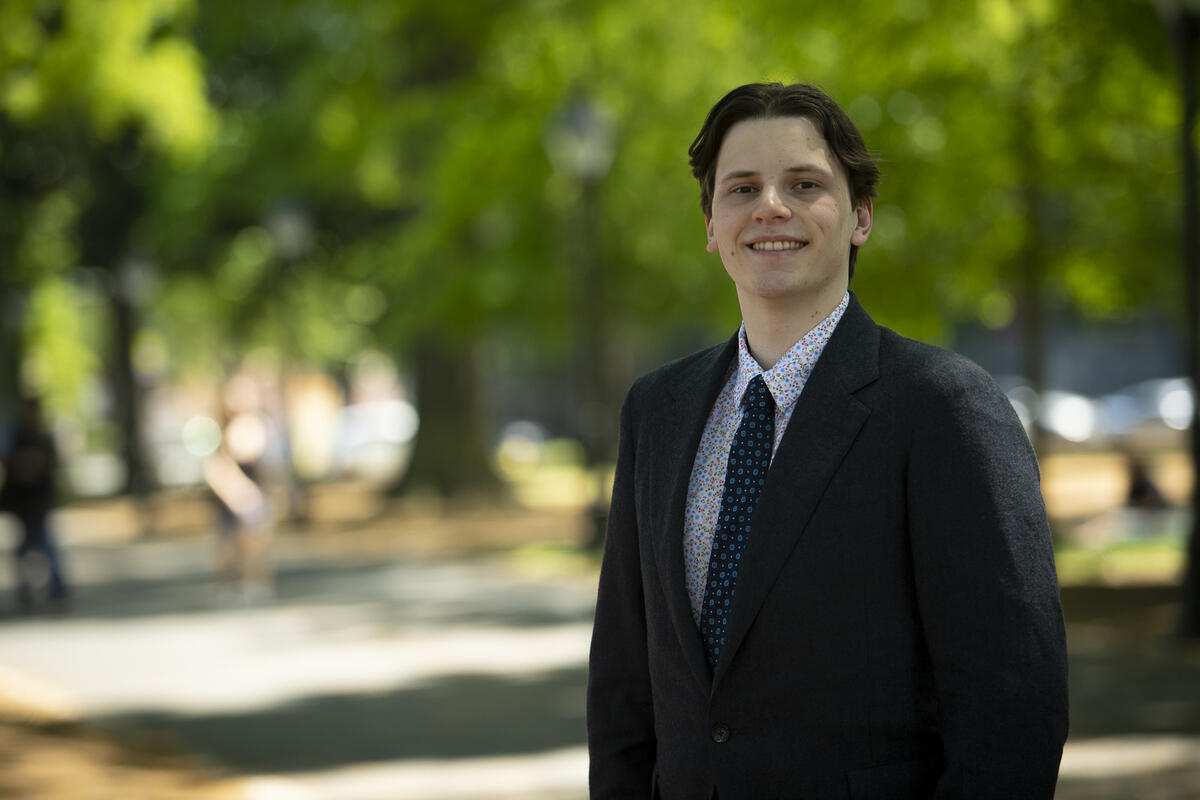 A man wearing a black suit jacket with a patterned tie and shirt standing in a park. 