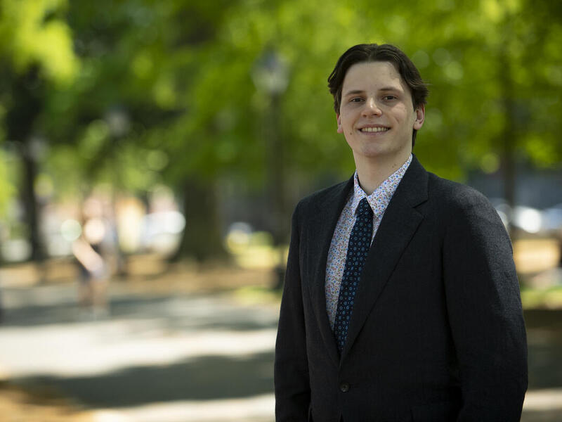 A man wearing a black suit jacket with a patterned tie and shirt standing in a park. 
