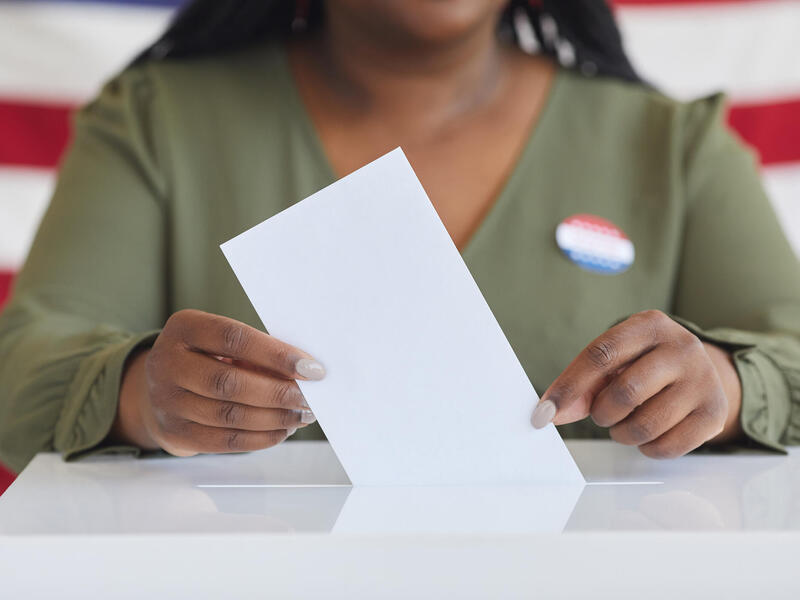 A photo of a woman's torso putting a piece of paper in a ballot box. 