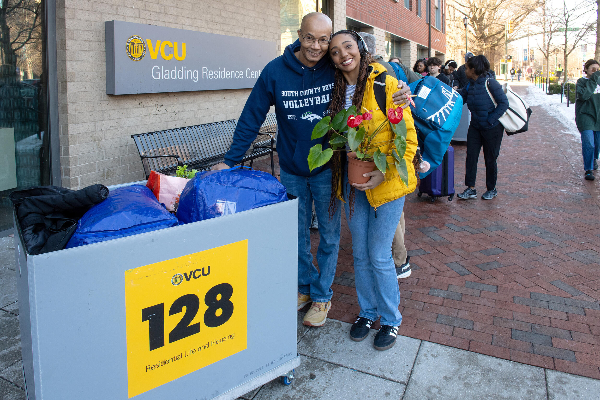 A photo of a man standing next to a young woman with his arm around her back. The woman is holding a potted plant in her right hand. There is a bin full of bags to the left of the man and woman. 