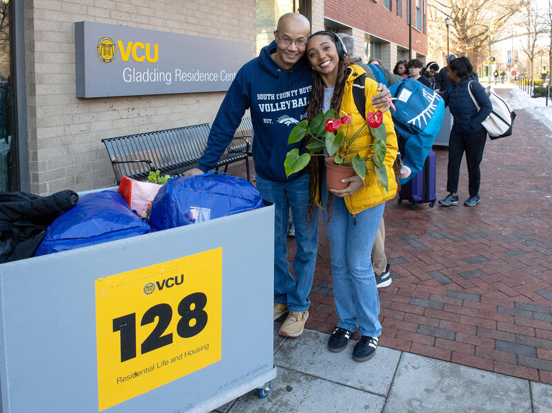 A photo of a man standing next to a young woman with his arm around her back. The woman is holding a potted plant in her right hand. There is a bin full of bags to the left of the man and woman. 