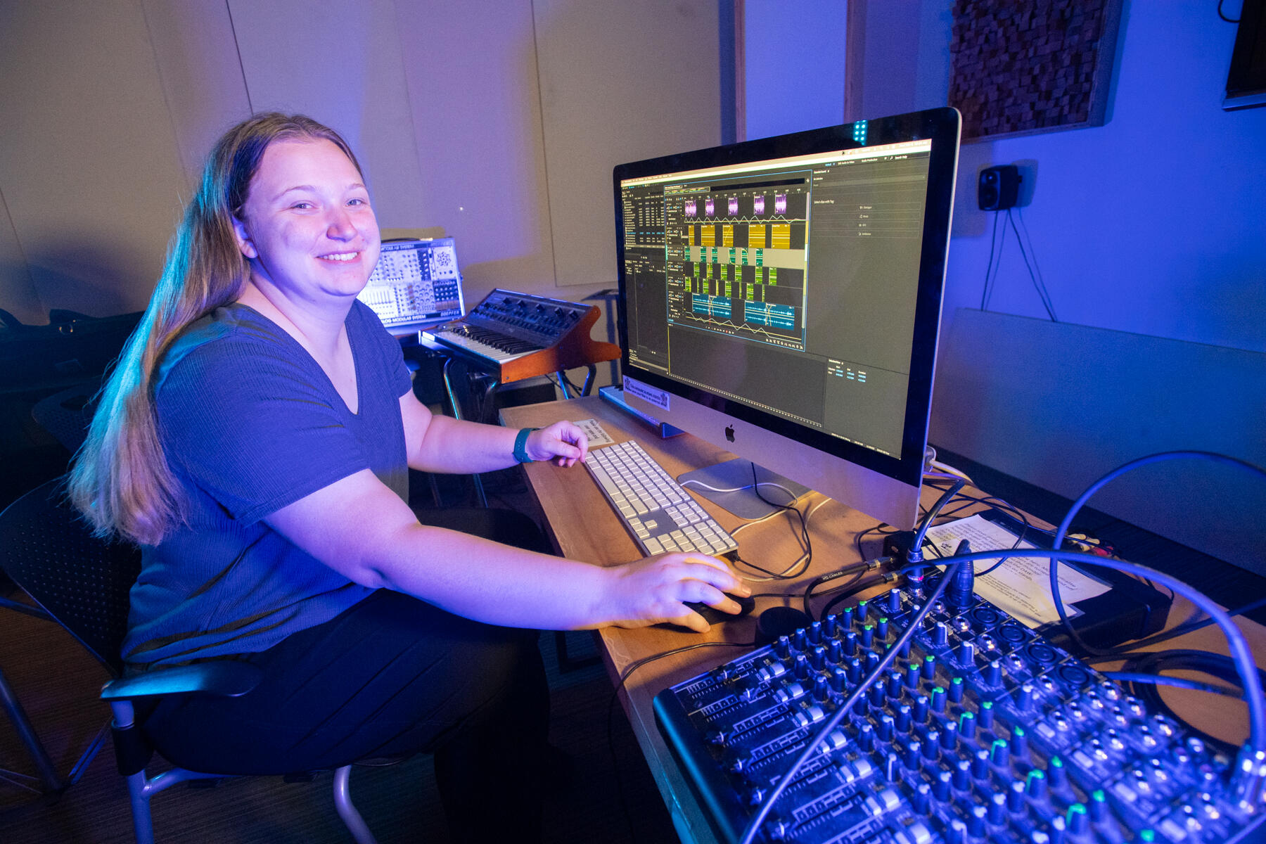 A photo of a woman sitting in front of a desk with a computer monitor on it. Next to the monitor on the desk is sound mixing equipment. 