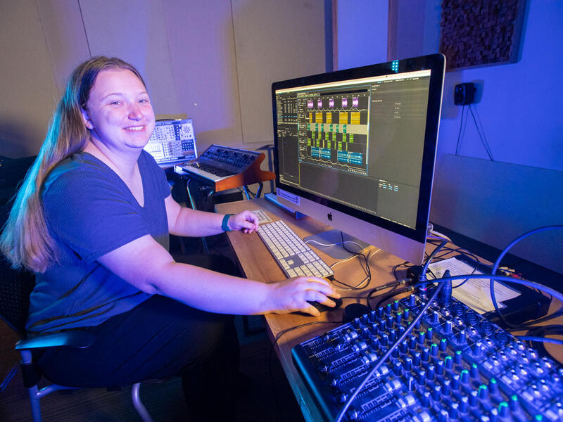 A photo of a woman sitting in front of a desk with a computer monitor on it. Next to the monitor on the desk is sound mixing equipment. 