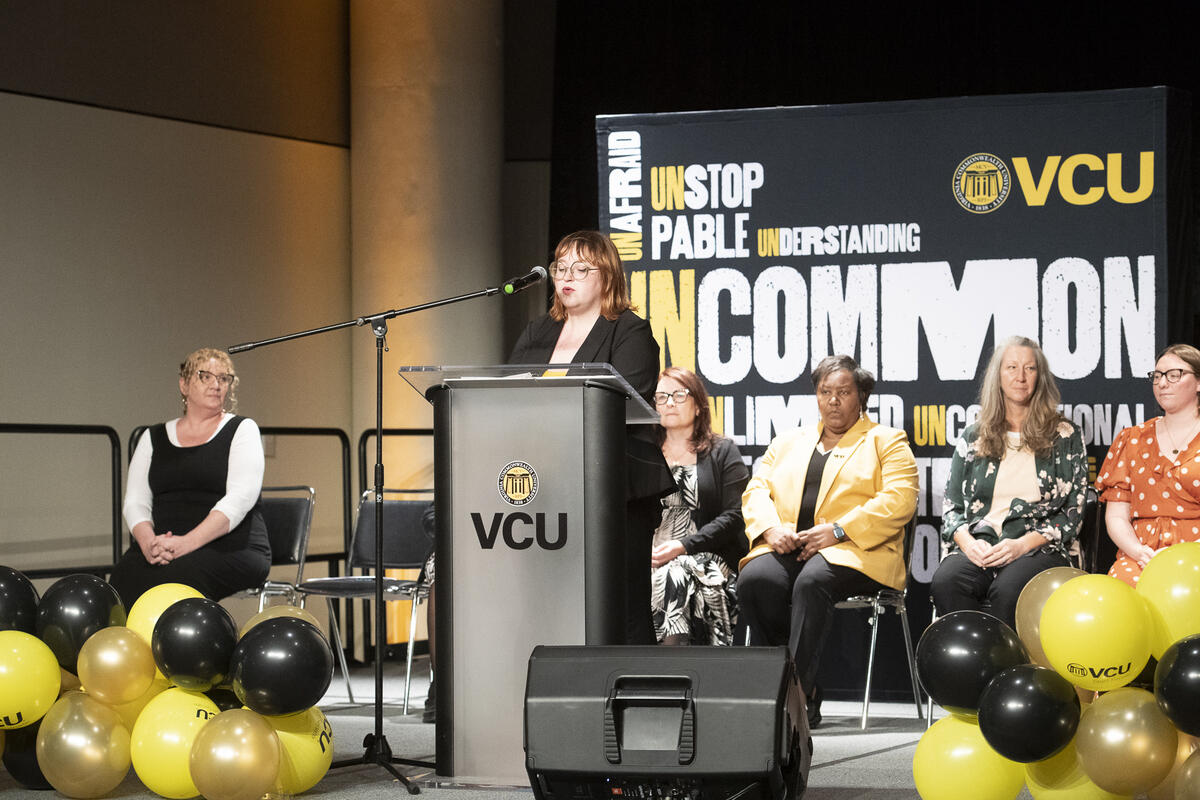 A woman standing at a podium and five other women seated behind her