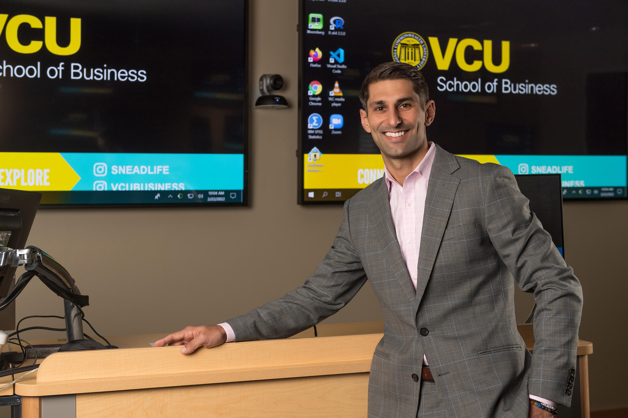 A photo of a man standing at the front of a lecture room with two screens behind him. 