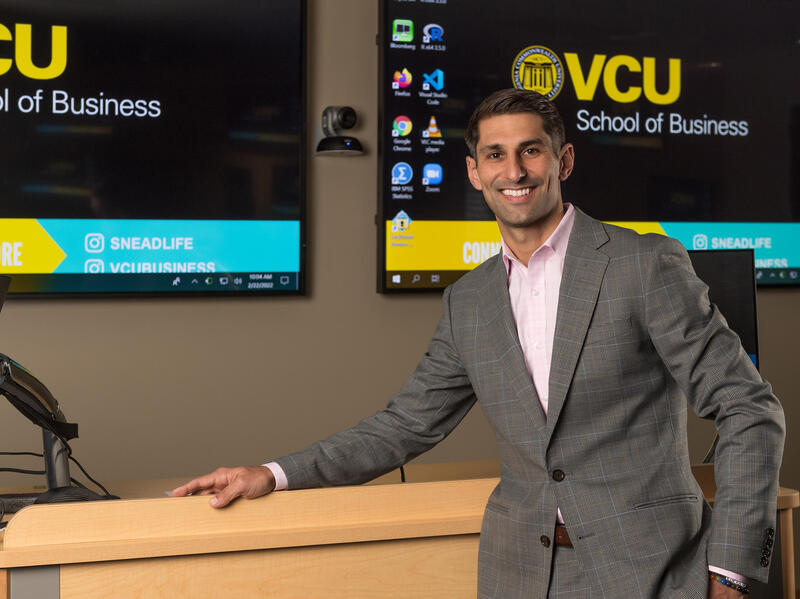A photo of a man standing at the front of a lecture room with two screens behind him. 