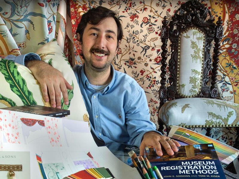 A photo of a man sitting with a pillow, illustrations and books. Behind him is vintage fabric and a vintage chair. 
