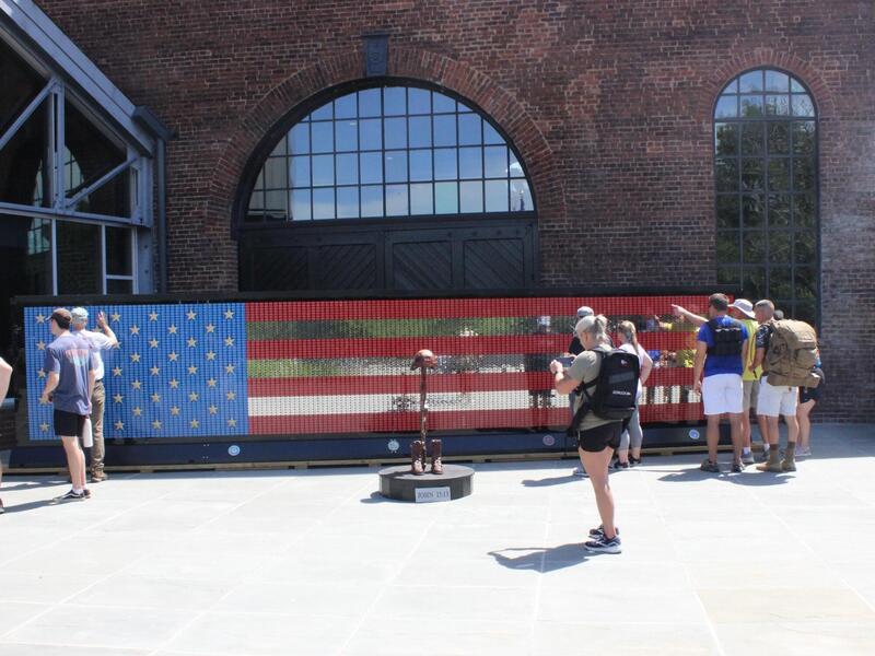 A photo of people standing in front of a large American flag made out of tiles. 