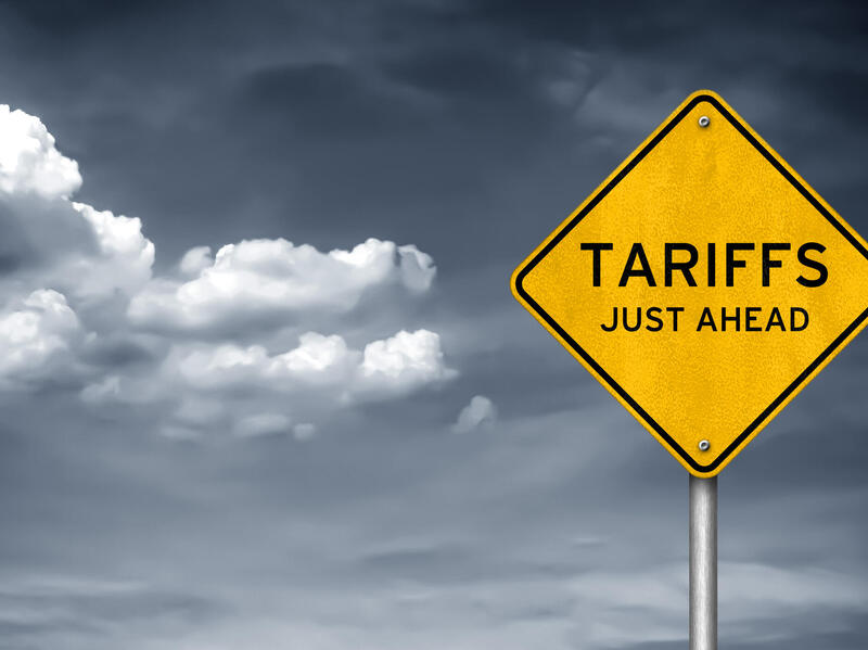 A photo of a yellow diamond shaped traffic sign that says \"TARIFFS JUST AHEAD\" in black letters. 