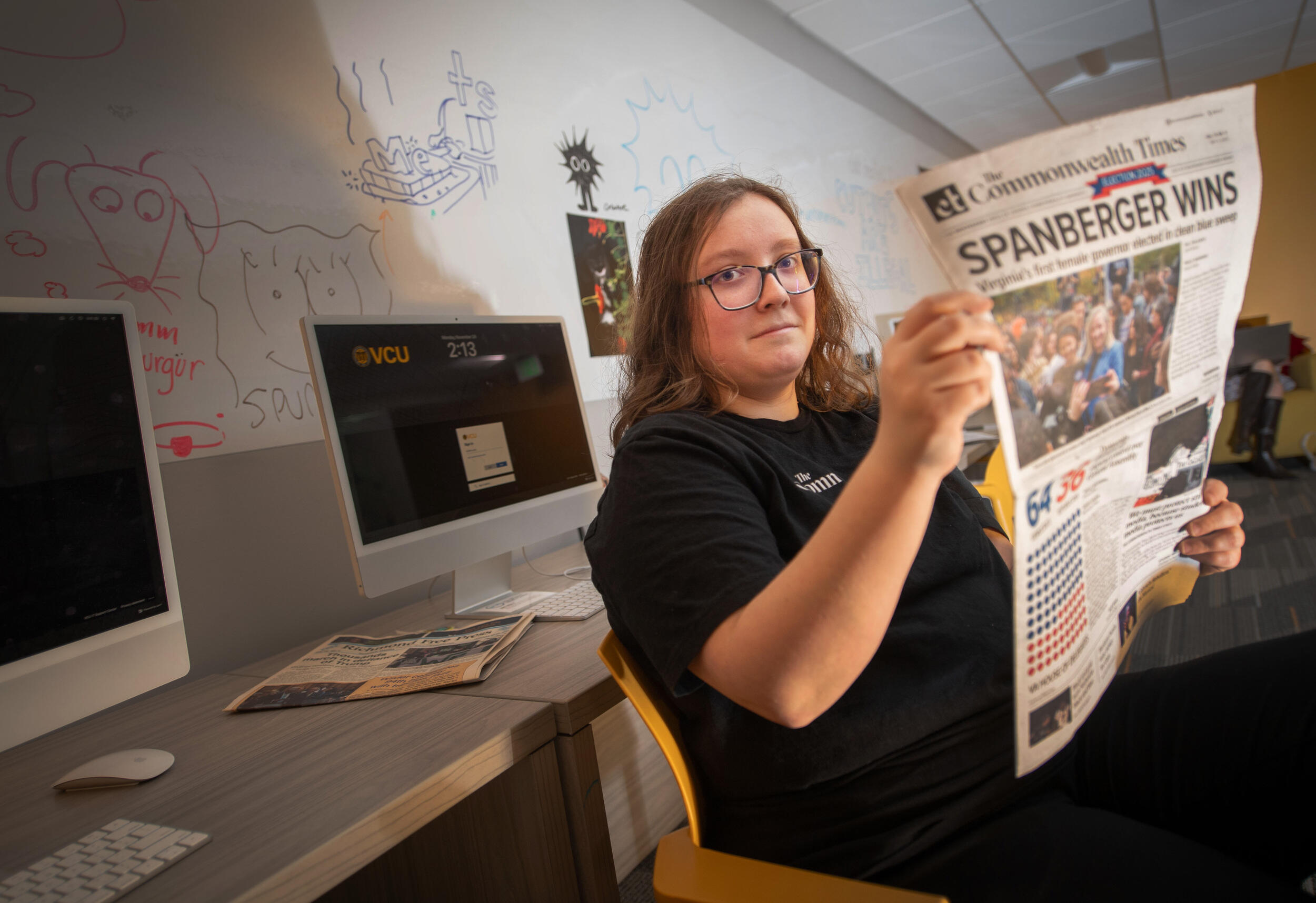 A photo of a woman sitting and holding a newspaper. Behind her is a whiteboard with illustrations drawn on it, and two computer monitors. 