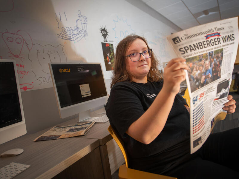 A photo of a woman sitting and holding a newspaper. Behind her is a whiteboard with illustrations drawn on it, and two computer monitors. 