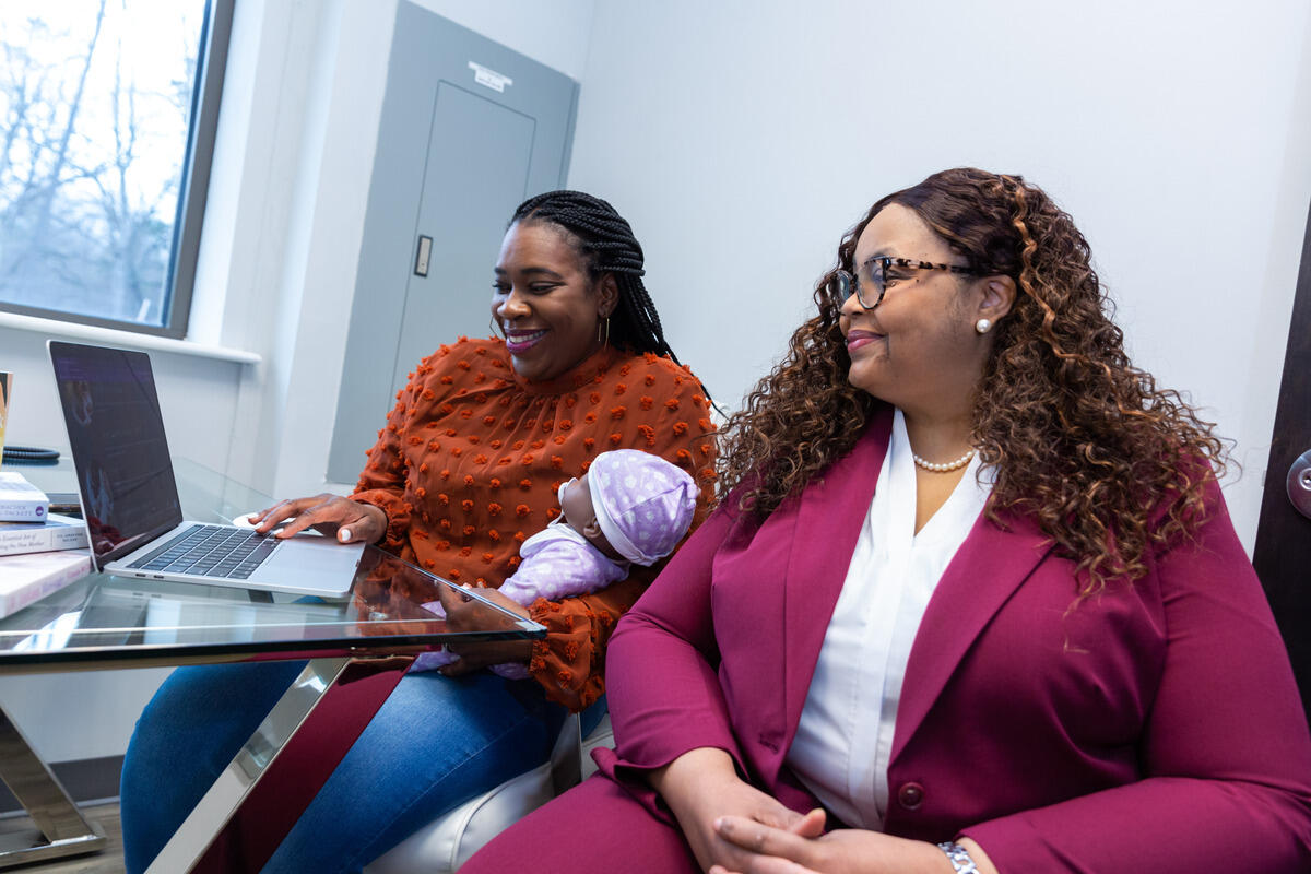 Two women sitting next to each other. The woman on the left is sitting at a table with a laptop and is holding a baby doll. 