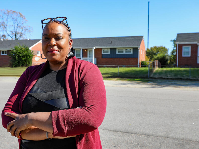 A photo of a woman from the waist up. Her arms are crossed against her chest. She is standing in the street in front of three houses. 
