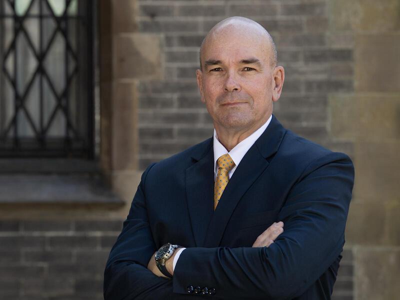 A photo of a bald man from the chest up. His arms are crossed against his chest. He is wearing a dark blue suit, a white button-up shirt, a yellow tie, and a black watch. 