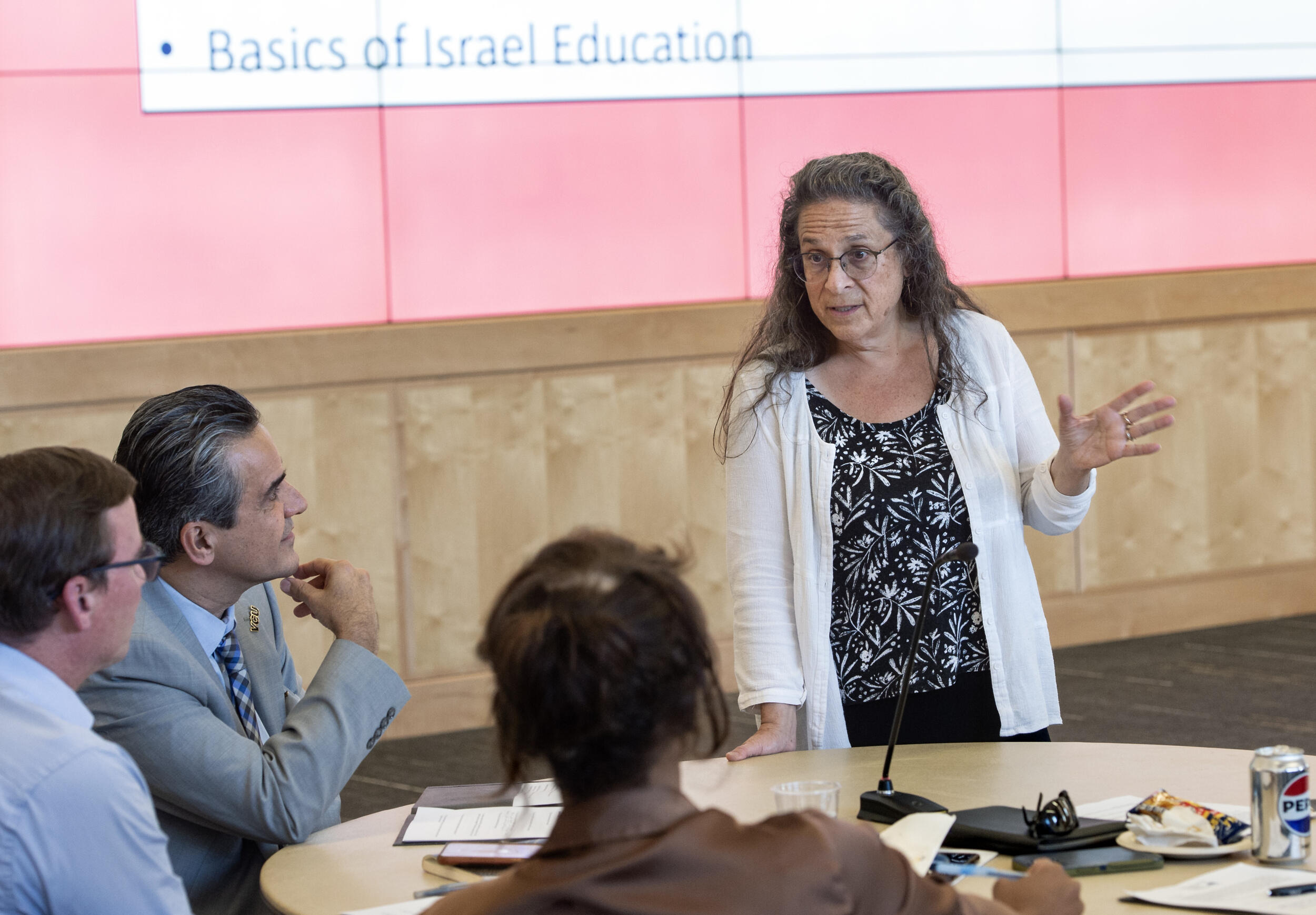 A photo of a woman standing in front of a circular table with three people sitting on the other side. She is speaking to the people at the table and they are looking at her. 