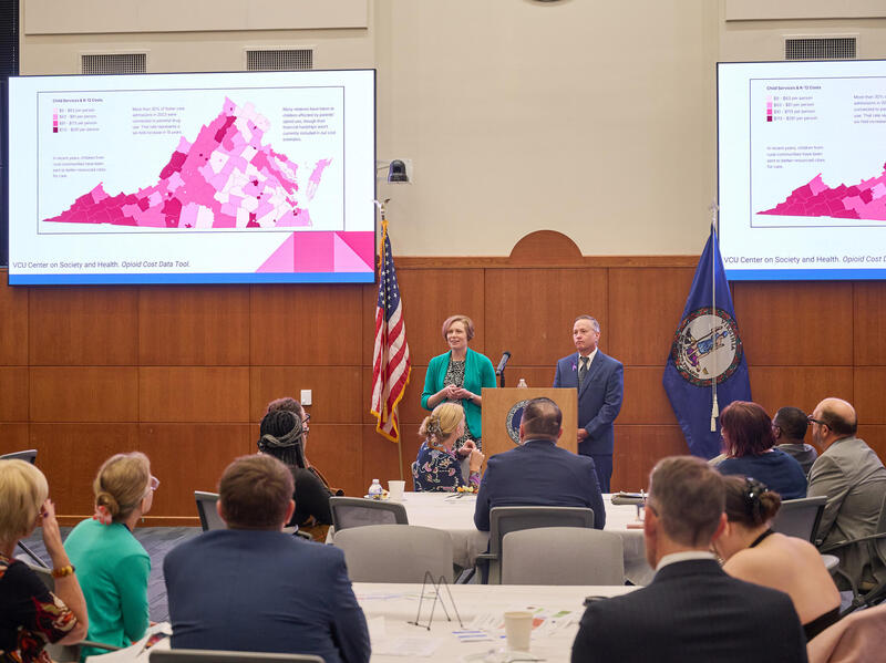 A photo of two people standing behind a podium in a room full iof people. Behind the two people presenting are two screens showing map of Virginia. 