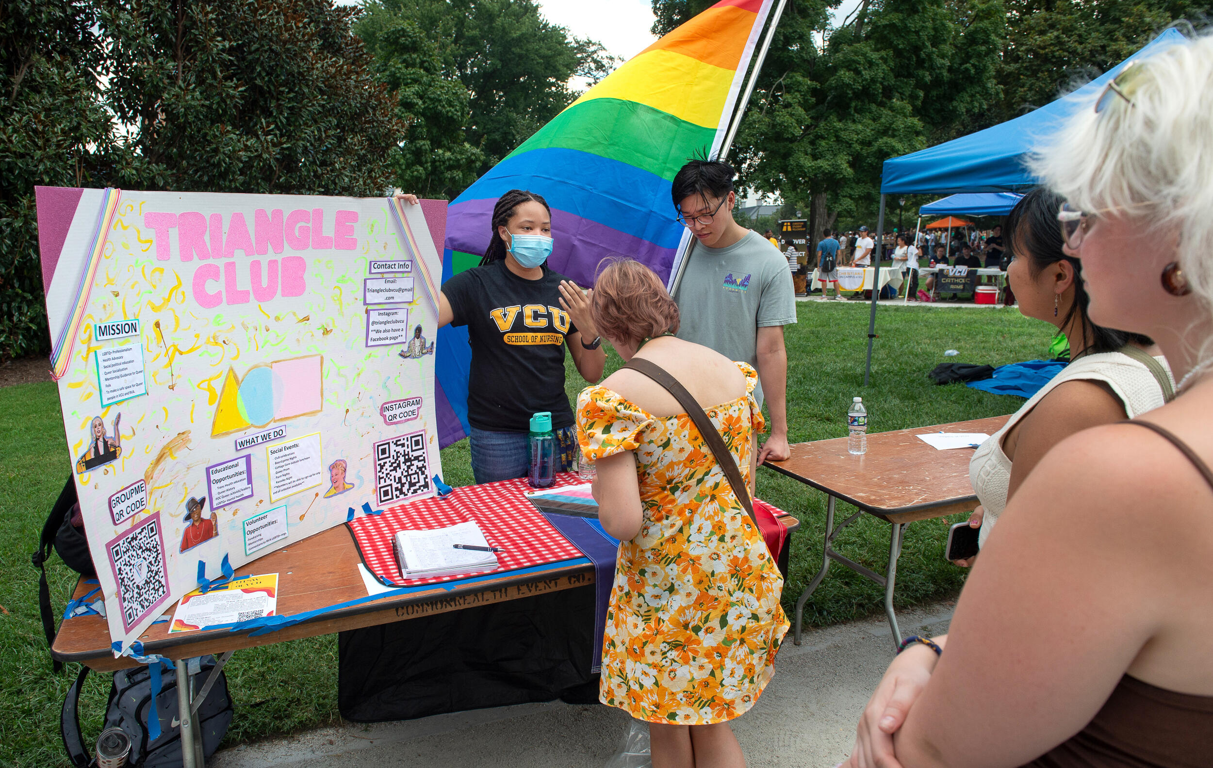 A student in front of a rainbow pride flag talks with another student at the Triangle Club booth of the Student Organization & Volunteer Opportunities Fair this fall along a sidewalk next to a tree-lined green space in Monroe Park.