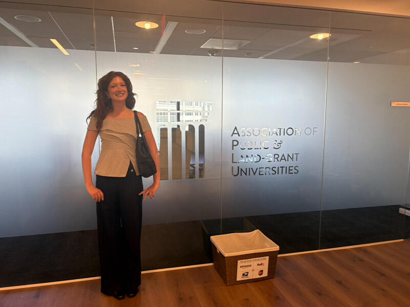 A photo of a woman standing in front of a glass wall. Written on the wall is the text \"Association of Public and Land-grant Universities.\" 