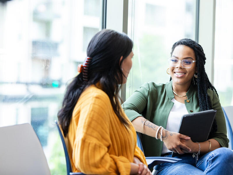 A photo of two women sitting in chairs talking to each other. The woman on the right is holding a clip board. 