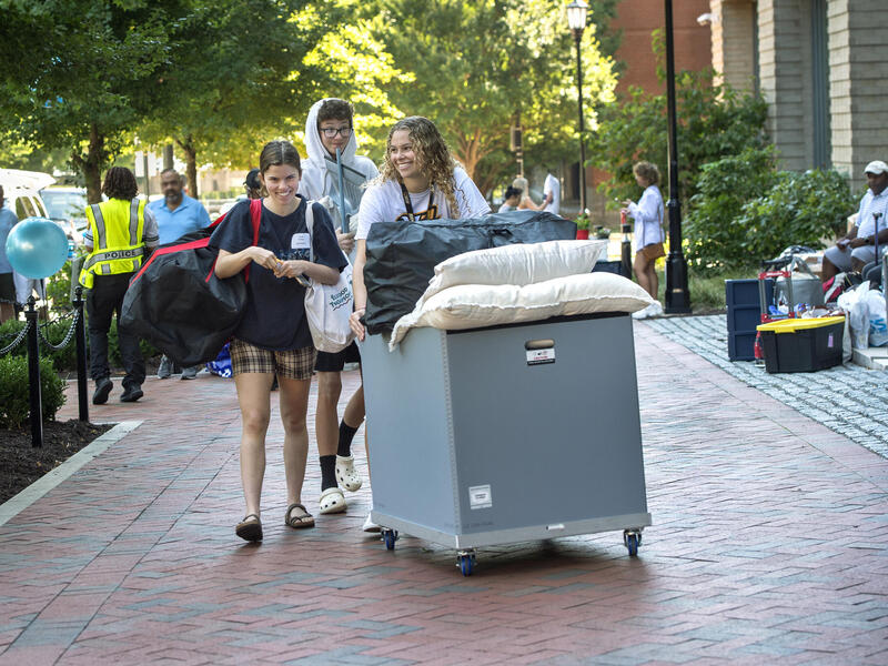 A photo of a student pushing a large gray bin on wheels down a brick sidewalk. Behind her is a young man wearing a hoodie, and to her right is a young woman carrying a large black bag with red straps over her shoulder. The three are smiling at each other. Behind them, parents and others are standing and sitting on the sides of the sidewalk. 