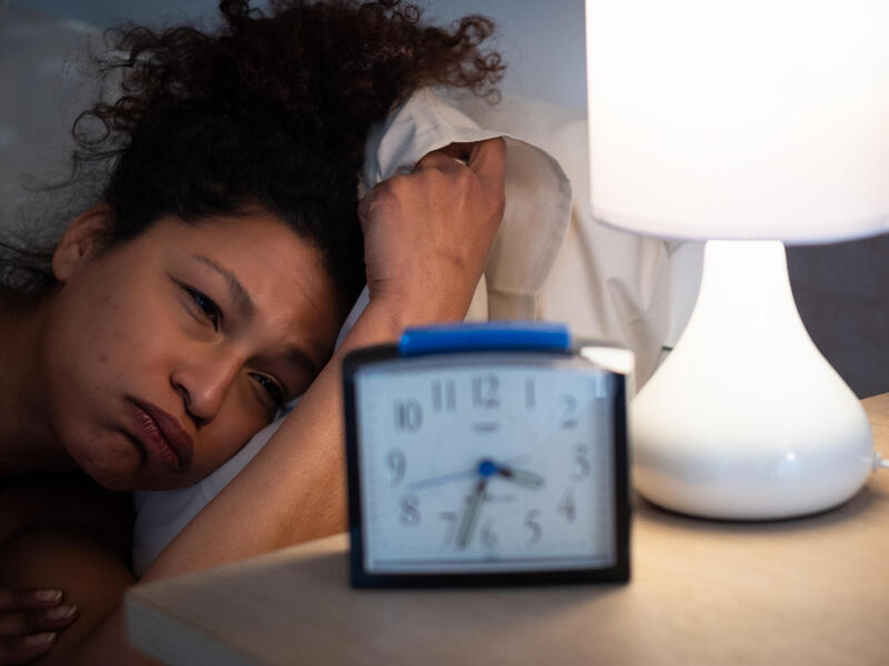 A woman laying in bed with a frustrated expression on her face as she stares at a clock next to her on a nightstand. 