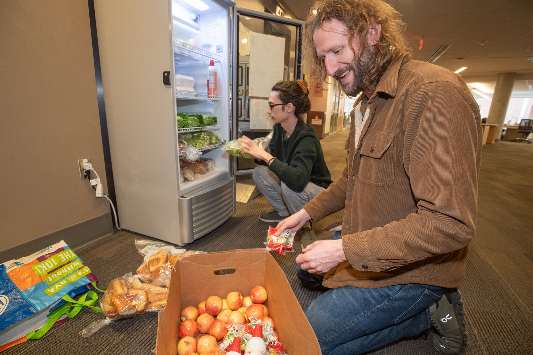 A photo of a man kneeling in front of a box of apples and another man kneeling in front of an open fridge. The man in front of the fridge is stocking it with produce. 