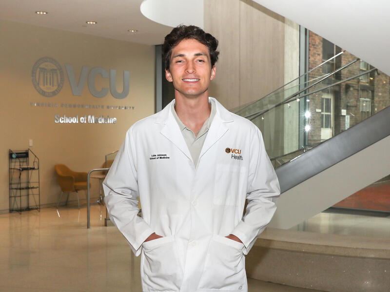 A photo of a man in a white medical coat from the waist up. He is smiling and his hands are in the two front pockets of the coat. 