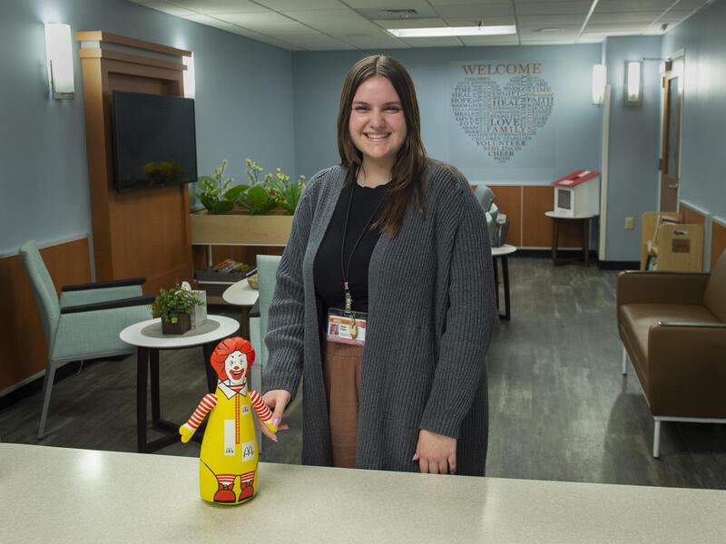 A woman standing in a room smiling. In front of her is a table with a red and yellow clown toy. 