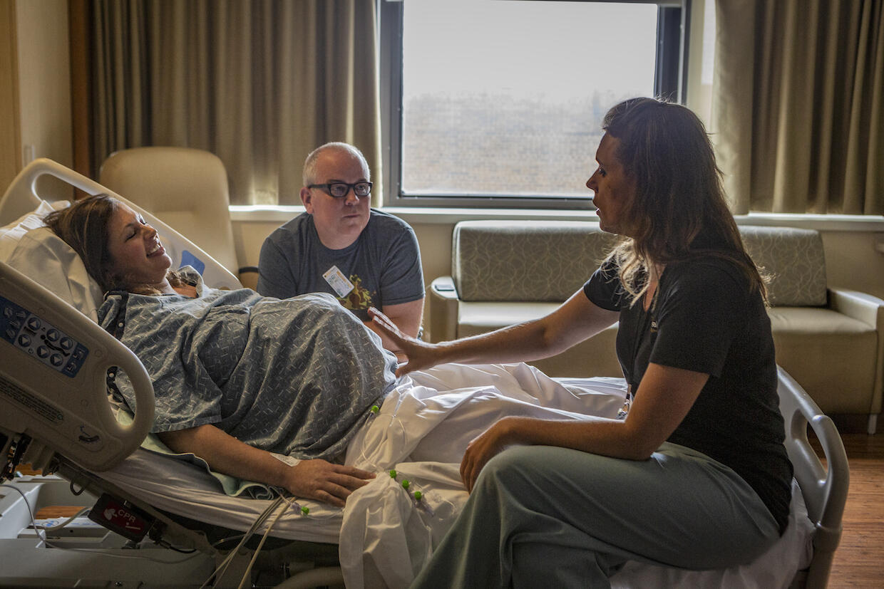 A smiling woman lying in a hospital bed smiles as a woman health care provider speaks with her. A man sitting next to the bed listens to the health care provider.