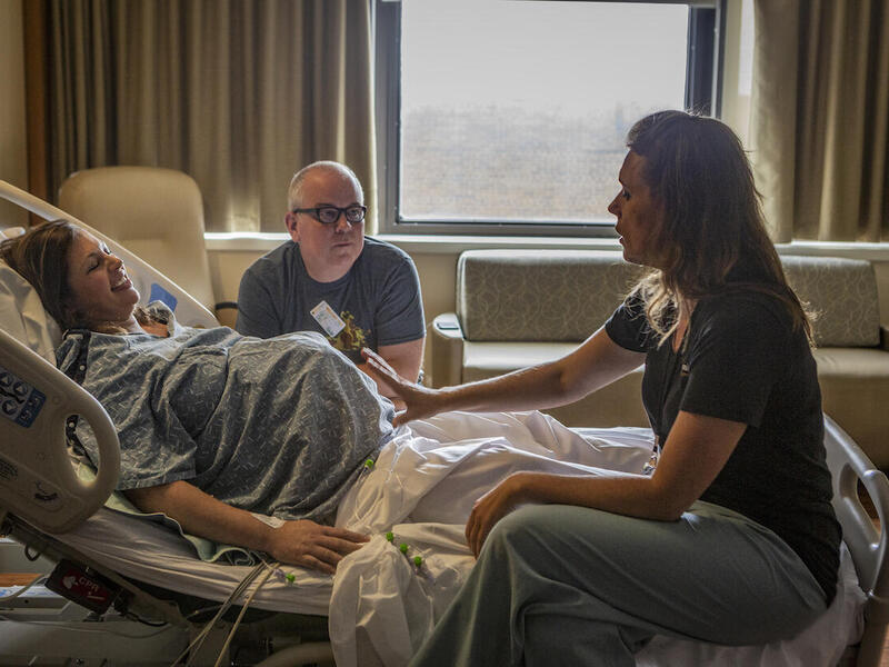 A smiling woman lying in a hospital bed smiles as a woman health care provider speaks with her. A man sitting next to the bed listens to the health care provider.