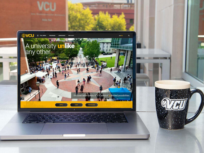A laptop sitting on a table in front of a window. Next to the laptop is a coffee mug that says \"VCU\" in white letters. 