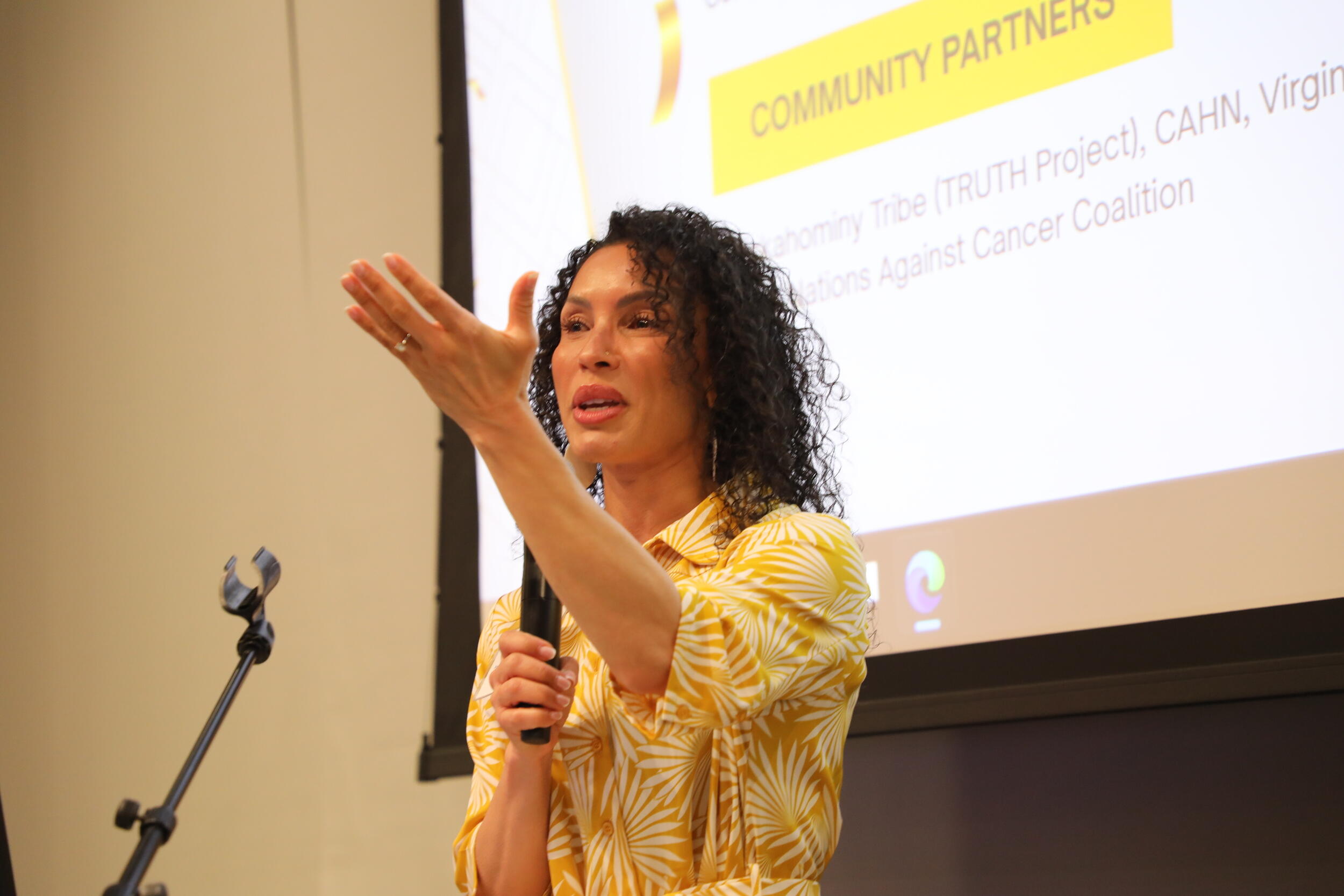 A photo of a woman from the waist up. She his holding a microphone and speaking while she gestures with her right hand. 