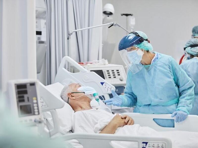 A photo of an elderly man laying in a hospital bed with a face mask on. Next to him stands a medical professional wearing a face mask, a face guard, latex gloves, and a disposable isolation gown. 
