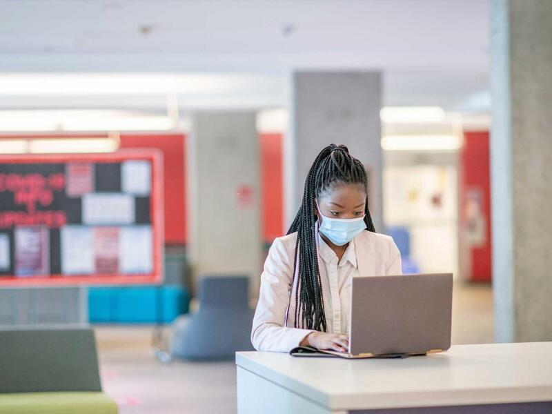 young black female college student sitting alone in a study space, wearing a face mask