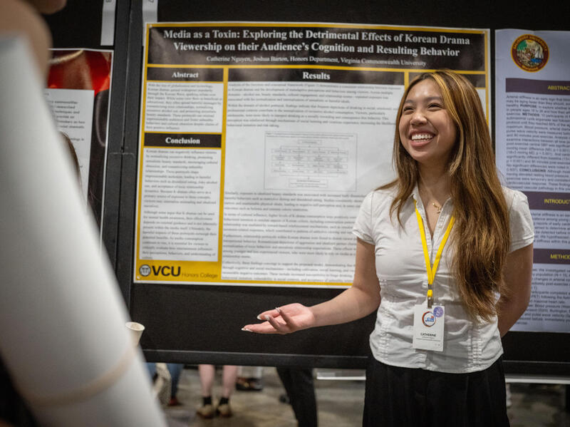 A photo of a woman standing in front of a research poster smiling and gesturing her left hand. 